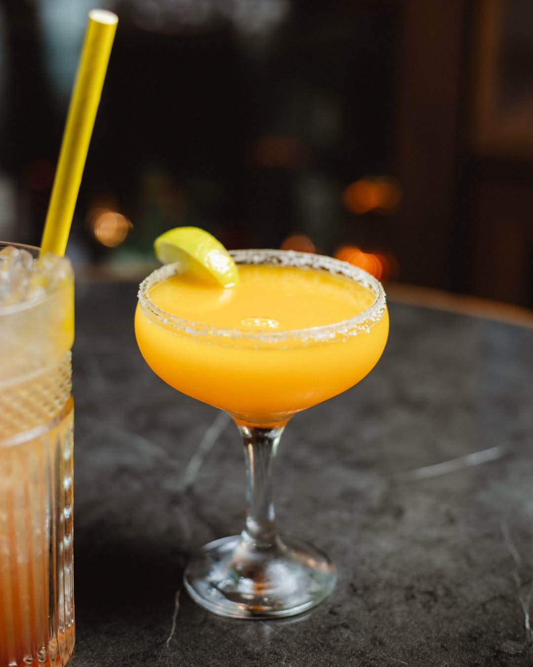 An amber cocktail in a sugar-rimmed coupe on the bar, lights low behind