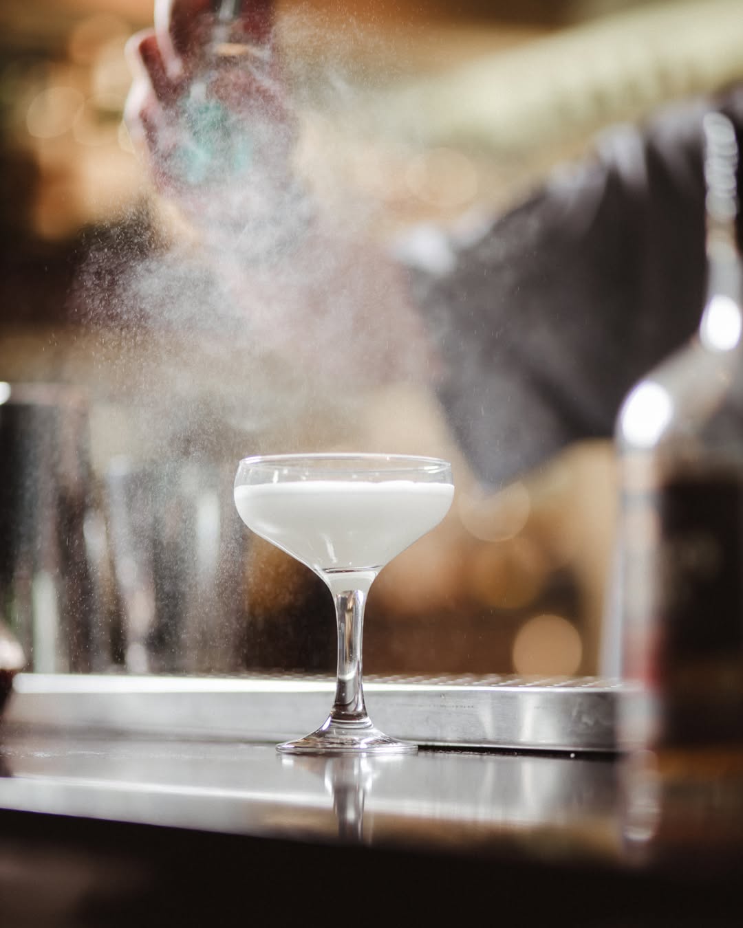 A white cocktail in a coupe caught in a cloud of aromatic mist at the bar