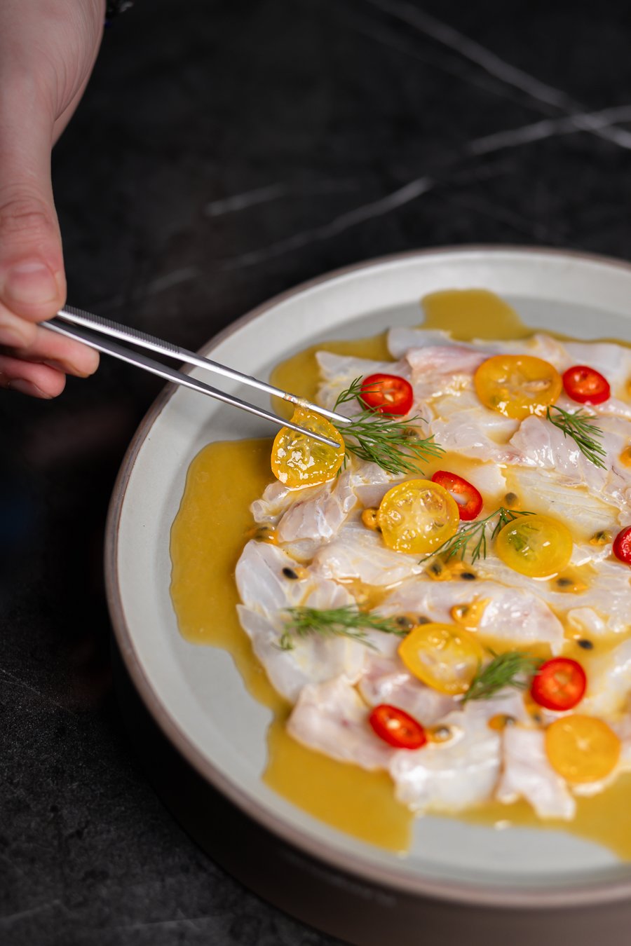 Sea bass carpaccio dressed with olive oil, yellow tomatoes, chili and dill, finished with tweezers at the pass