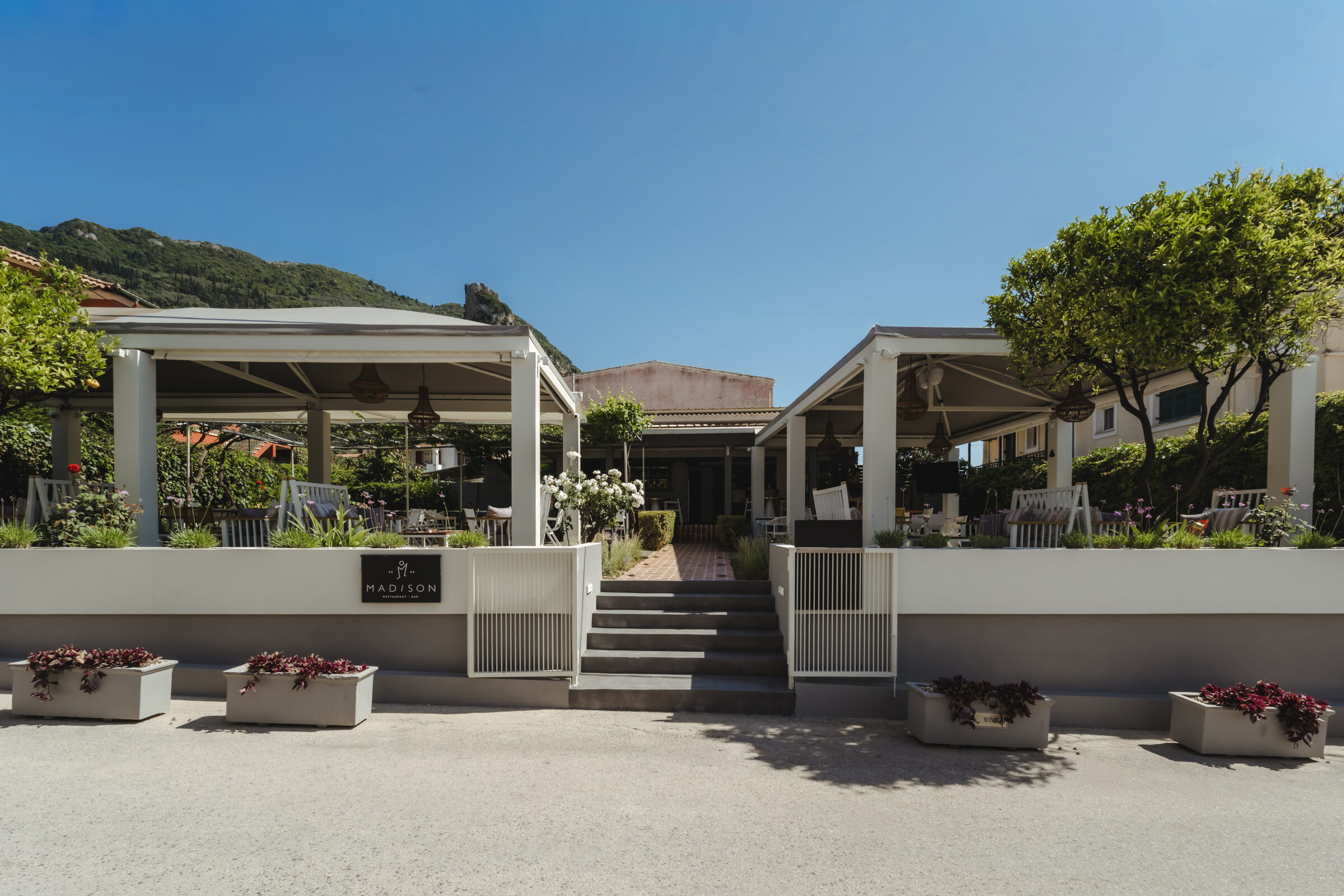 The entrance to Madison Garden in daylight, framed by citrus trees and the Corfu hills