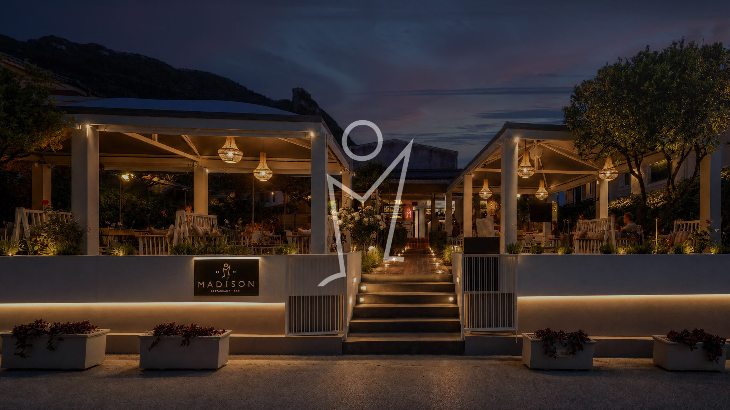 Madison Garden at dusk, terrace lit with warm lanterns beneath the Agios Gordios hillside